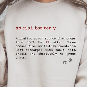 May include: Cream-colored sweatshirt with the text "social battery" in red, followed by a definition. The definition describes a limited power source that drops after small talk, recharged by books, pets, and snacks. Includes paw prints.