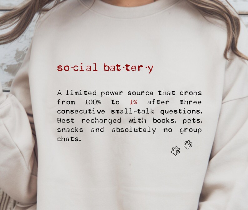 May include: Cream-colored sweatshirt with the text "social battery" in red, followed by a definition. The definition describes a limited power source that drops after small talk, recharged by books, pets, and snacks. Includes paw prints.
