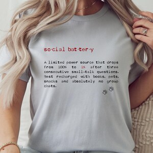 May include: Light gray t-shirt with the text "social battery" in red and black. The text describes a limited power source that drops from 100% to 1% after small talk, recharged by books, pets, and snacks.
