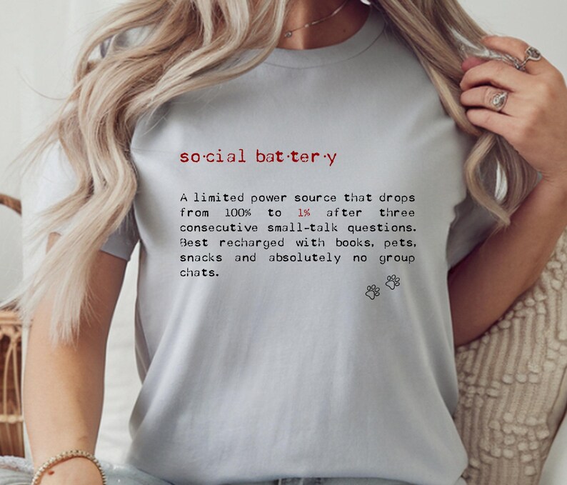 May include: Light gray t-shirt with the text "social battery" in red and black. The text describes a limited power source that drops from 100% to 1% after small talk, recharged by books, pets, and snacks.