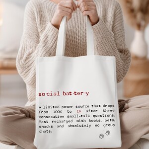 May include: White tote bag with the words "social battery" in red and black text. The text describes a limited power source that drops from 100% to 1% after small talk, recharged by books, pets, and snacks.