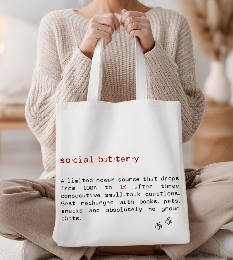 May include: White tote bag with the words "social battery" in red and black text. The text describes a limited power source that drops from 100% to 1% after small talk, recharged by books, pets, and snacks.