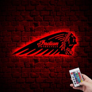 Puede incluir: Una luz LED roja con el logotipo de Indian Motorcycle. El logotipo presenta una cabeza de nativo americano estilizada con un tocado de plumas y las palabras "Indian Motorcycle". La luz está montada en una pared de ladrillo y se controla mediante un mando a distancia con múltiples opciones de color.