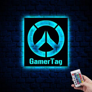 以下が含まれることがあります： 「GamerTag」という文字とスタイリッシュな平和のシンボルが書かれた黒と青の光る看板。看板はレンガの壁に取り付けられており、青い光で照らされています。画像の右下には、複数の色のボタンが付いたリモコンが見えます。
