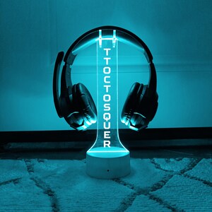 Puede incluir: Un soporte de luz LED azul con la palabra "TOCTOSQUER" impresa en él. Un par de auriculares negros están apoyados en el soporte.