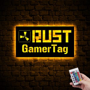 May include: A black rectangular sign with yellow LED lights around the border. The sign says "RUST Gamer Tag" with a stylized symbol of a radiation symbol to the left of the text. A remote control with colour changing buttons is visible in the bottom right corner of the image.