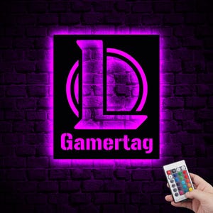 Puede incluir: Un letrero de ne&oacute;n con un brillo morado, que presenta una letra "L" estilizada dentro de un c&iacute;rculo, y el texto "Gamertag" debajo. El letrero est&aacute; montado en una pared de ladrillo e incluye un control remoto con opciones de color.