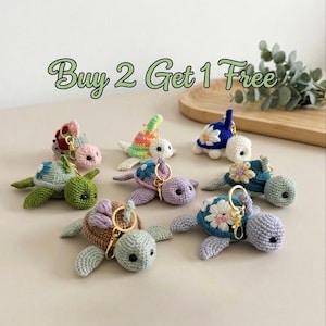 Può includere: Una collezione di portachiavi a forma di tartaruga fatti a mano all'uncinetto in vari colori, tra cui verde, blu, viola e marrone. Ogni tartaruga presenta un design unico con decorazioni floreali e un anello portachiavi dorato. Il testo "Buy 2 Get 1 Free" è visibile.