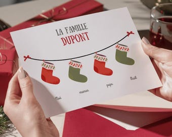 May include: A Christmas card held by hands, featuring the text "LA FAMILLE DUPONT" in red. Below, four Christmas stockings hang from a string, each with a name: Noé, maman, papa, and Maël. The stockings are red and green.