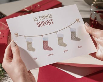 May include: A greeting card with the text "LA FAMILLE DUPONT" in burgundy. The card features illustrations of Christmas stockings hanging from a string, each labeled with a family member's name. The card is held by a person.