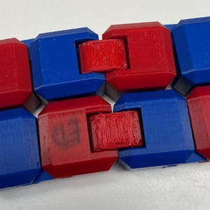 Könnte beinhalten: Ein rotes und blaues Fidget-Spielzeug mit quadratischer Form und einem Drehmechanismus. Das Spielzeug ist aus Kunststoff und hat eine glatte Oberfläche.