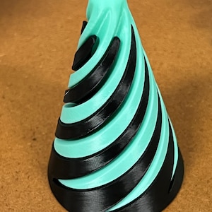 Op de afbeelding: Een turquoise kegel met zwarte spiraalvormige uitsnijdingen. De kegel is gemaakt van plastic en heeft een glad oppervlak.