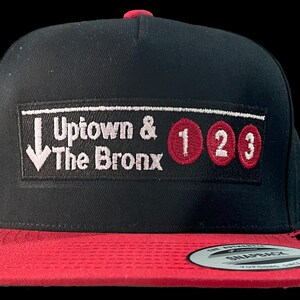 Puede incluir: Gorra de béisbol negra y roja con un parche bordado que dice "Uptown & The Bronx" con los números 1, 2 y 3 en círculos rojos.