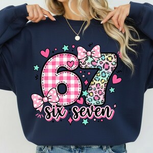May include: Navy blue sweatshirt with a colorful graphic design. The design features the numbers "67" with a pink gingham pattern for the "6" and a leopard print for the "7". The words "six seven" are written below the numbers in a script font.