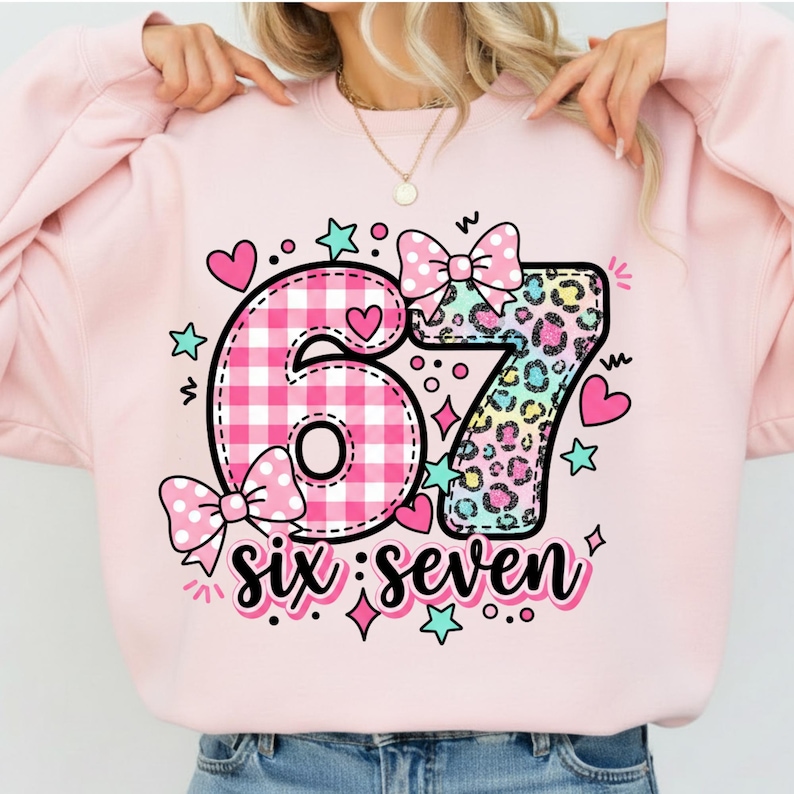 May include: Pink sweatshirt with a colorful graphic design. The design features the numbers "67" with a gingham pattern and leopard print, along with the words "six: seven" and decorative hearts, stars, and bows. A fun and festive design.
