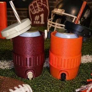 May include: Two insulated drink coolers, one maroon and one orange, with lids and straws, sit on a green field. A football and pom-poms are in the foreground. The background includes a football play diagram and a sign that says "GAME DAY."