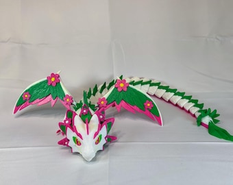 Könnte beinhalten: Eine weiße Drachenfigur mit grünen Flügeln und einem rosa Schwanz. Der Drache ist mit rosa Blumen verziert und hat ein weißes Gesicht mit grünen Augen.
