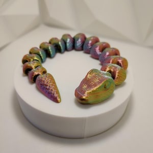 Op de afbeelding: Een regenboogkleurige, slangvormige armband van plastic. De armband bestaat uit afzonderlijke segmenten die aan elkaar zijn verbonden. De armband ligt op een wit oppervlak.