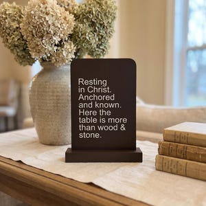 Può includere: Un cartello di legno marrone scuro con il testo "Resting in Christ. Anchored and known. Here the table is more than wood & stone." Un vaso di fiori secchi è dietro il cartello. Tre vecchi libri sono sulla destra.