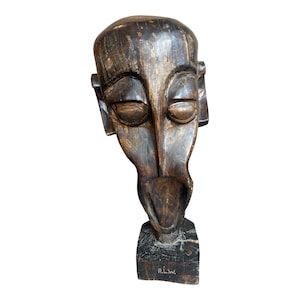 May include: A dark brown, carved wooden sculpture of a stylized face. The sculpture has closed eyes, a long nose, and a protruding lower lip. The base is rectangular, and the initials "R.L.W." are carved on the front.
