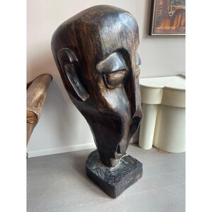 May include: A large, dark brown wooden sculpture of a human head with closed eyes and a prominent nose. The sculpture is mounted on a square base and has a rough, textured surface. The artwork is a unique piece of home decor.