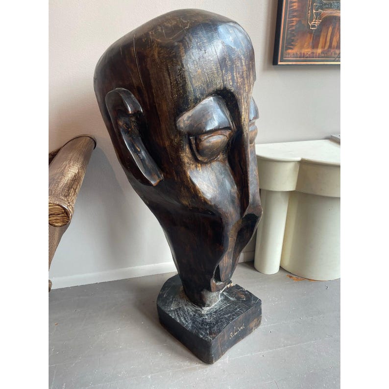 May include: A large, dark brown wooden sculpture of a human head with closed eyes and a prominent nose. The sculpture is mounted on a square base and has a rough, textured surface. The artwork is a unique piece of home decor.