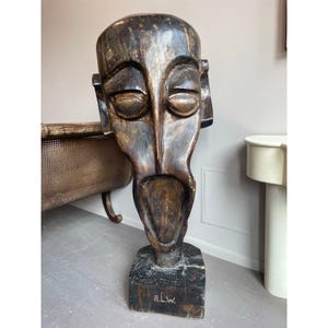 May include: A large, carved wooden mask with a stylized human face. The mask is primarily dark brown with lighter highlights, and features a wide open mouth and prominent facial features. The base is a dark square with the initials "R.L.W." carved into it.