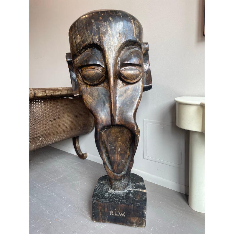 May include: A large, carved wooden mask with a stylized human face. The mask is primarily dark brown with lighter highlights, and features a wide open mouth and prominent facial features. The base is a dark square with the initials "R.L.W." carved into it.