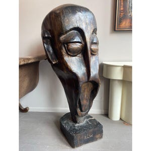 May include: A large, dark brown wooden sculpture of a stylized human head with exaggerated features. The sculpture has a rough, textured surface and is mounted on a square base. The artist's initials, "R.L.W.", are carved into the base.