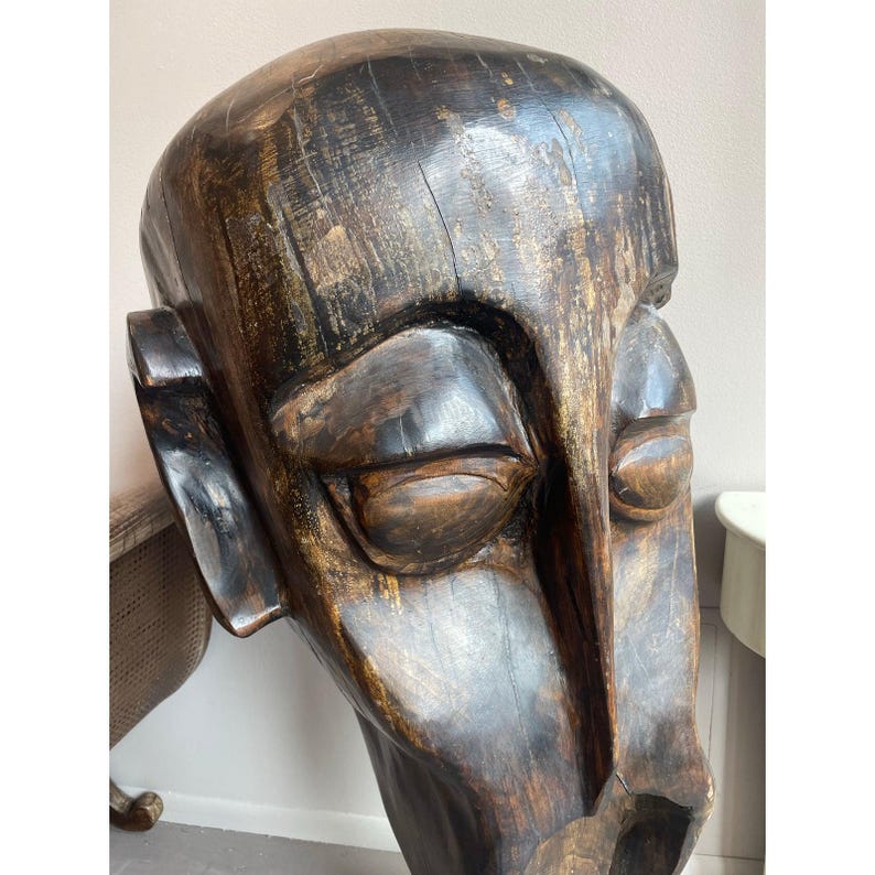 May include: A large, carved wooden sculpture of a human head. The sculpture is dark brown with visible wood grain and a smooth, polished finish. The face has closed eyes, a prominent nose, and a slightly open mouth, suggesting a contemplative expression.