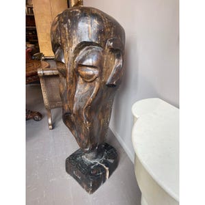 May include: A large, carved wooden sculpture of a human head. The sculpture is dark brown with visible wood grain and features carved facial details. It stands on a dark, square base. The artwork is a unique piece of home decor.