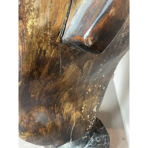 May include: Close-up of a wooden sculpture with a dark brown finish and gold accents. The sculpture has a rough, textured surface with visible cracks and imperfections. The base appears to be made of a dark, possibly marble-like material.