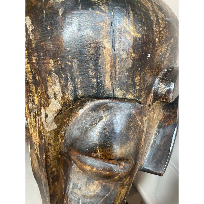 May include: Close-up of a carved wooden sculpture of a face. The sculpture is primarily dark brown with lighter brown and gold accents. The face has a prominent nose, closed lips, and a defined jawline. The wood grain is visible.