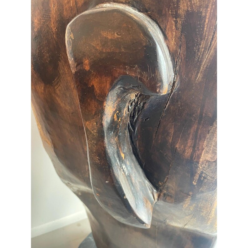 May include: Close-up of a carved wooden sculpture, featuring a detailed ear and part of a face. The wood has a rich, dark brown color with visible grain and texture, suggesting a natural, handcrafted piece. The sculpture appears to be part of a larger artwork.