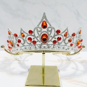 May include: A silver-toned metal tiara featuring a symmetrical, ornate scrollwork design encrusted with numerous small clear rhinestones and accented with multiple vibrant orange, teardrop-shaped and round faceted crystals throughout the piece.