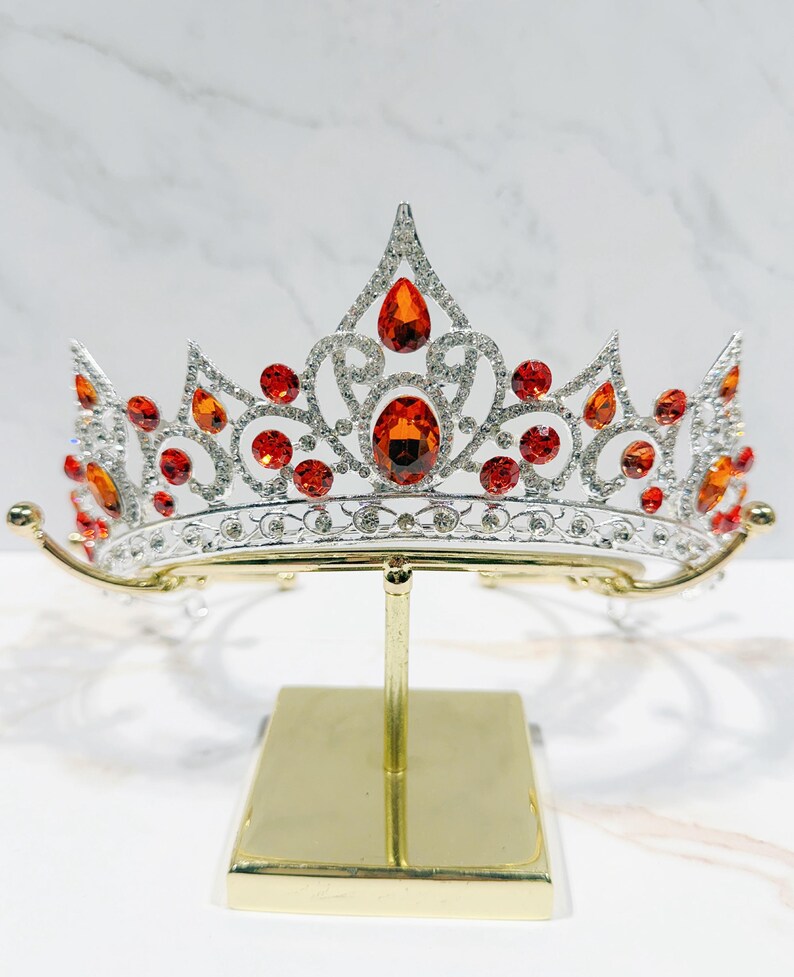 May include: A silver-toned metal tiara featuring a symmetrical, ornate scrollwork design encrusted with numerous small clear rhinestones and accented with multiple vibrant orange, teardrop-shaped and round faceted crystals throughout the piece.