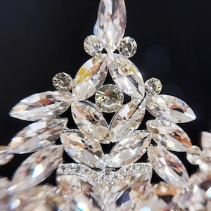 May include: Close-up of a sparkling tiara with clear, faceted crystals. The tiara features a central, elongated crystal surrounded by smaller, round and marquise-shaped stones. The design is symmetrical and elegant, perfect for special occasions.