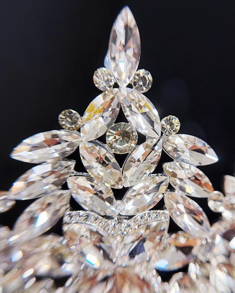 May include: Close-up of a sparkling tiara with clear, faceted crystals. The tiara features a central, elongated crystal surrounded by smaller, round and marquise-shaped stones. The design is symmetrical and elegant, perfect for special occasions.