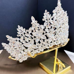 May include: A silver-colored crown with clear, sparkling crystals arranged in a floral pattern. The crown is displayed on a gold-colored stand with a square base. The crystals reflect light, creating a dazzling effect. The crown is likely for a special occasion.