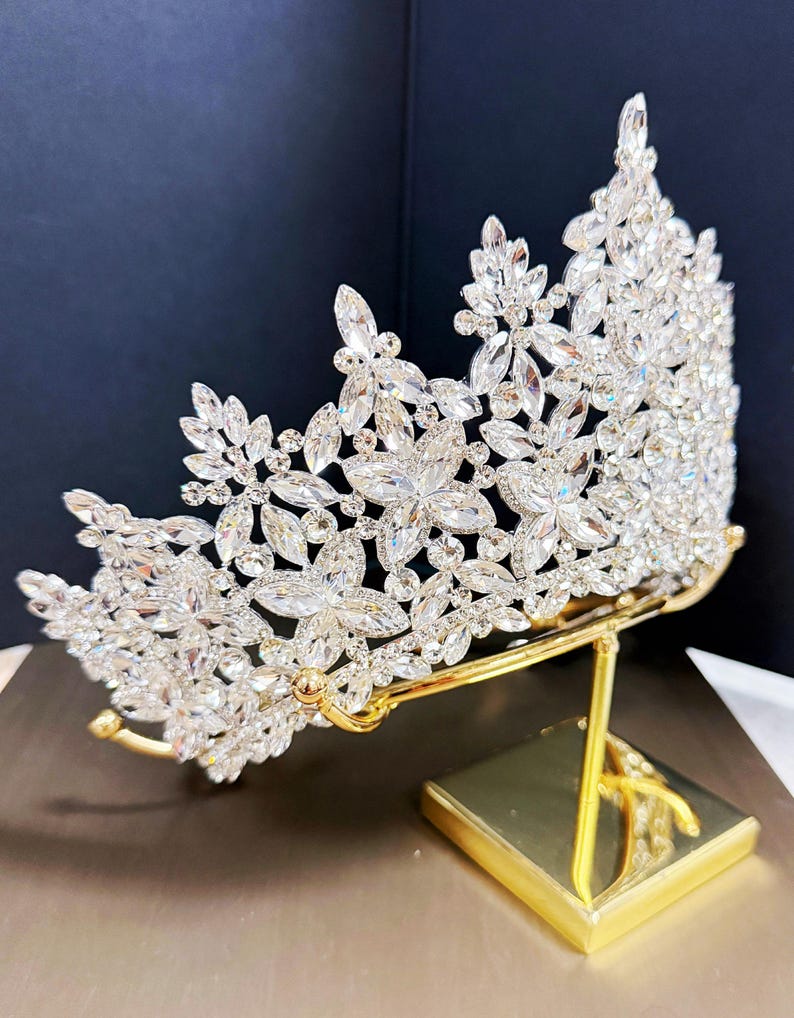 May include: A silver-colored crown with clear, sparkling crystals arranged in a floral pattern. The crown is displayed on a gold-colored stand with a square base. The crystals reflect light, creating a dazzling effect. The crown is likely for a special occasion.