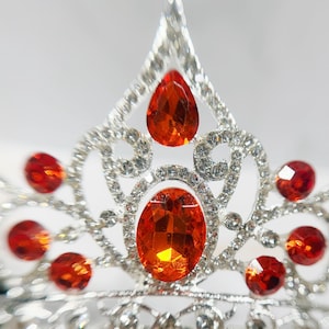 May include: A silver-toned tiara featuring a central oval-shaped orange gemstone, a pear-shaped orange gemstone at the peak, and multiple smaller round orange gemstones set within a decorative metal frame encrusted with clear crystals.