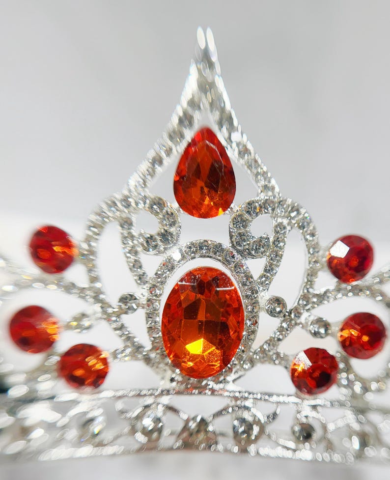 May include: A silver-toned tiara featuring a central oval-shaped orange gemstone, a pear-shaped orange gemstone at the peak, and multiple smaller round orange gemstones set within a decorative metal frame encrusted with clear crystals.