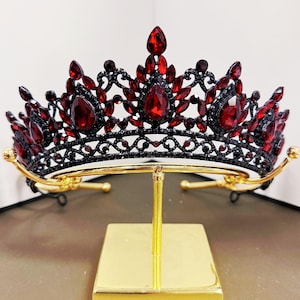 May include: A black metal tiara featuring intricate filigree scrollwork and numerous deep red teardrop and marquise-cut crystals. The crown is adorned with smaller black rhinestones throughout the frame, creating a dark, ornate appearance.