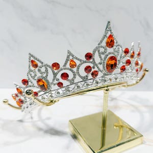 May include: A silver-toned metal tiara featuring intricate scrollwork patterns and numerous clear rhinestones. The headpiece is accented with multiple orange and red teardrop-shaped and round crystals, displayed on a gold-colored metal stand.