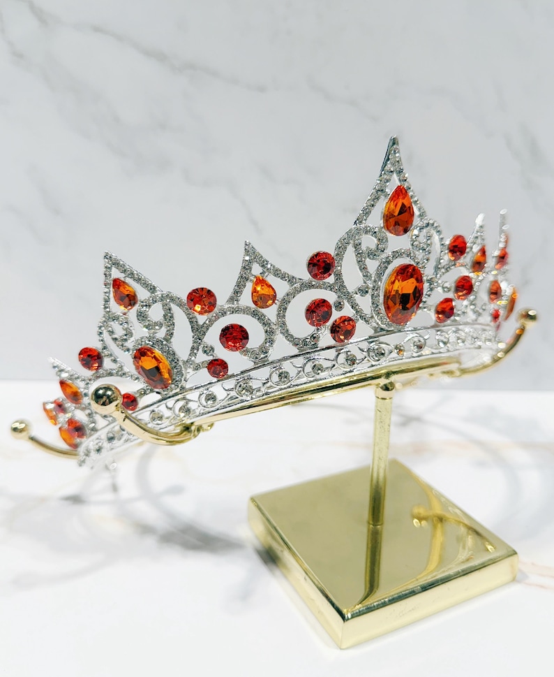 May include: A silver-toned metal tiara featuring intricate scrollwork patterns and numerous clear rhinestones. The headpiece is accented with multiple orange and red teardrop-shaped and round crystals, displayed on a gold-colored metal stand.