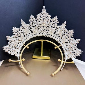 May include: A silver-toned crown with a floral design, adorned with clear crystals. The crown features a gold-colored base with a curved design and small, round accents. The piece is designed for special occasions.