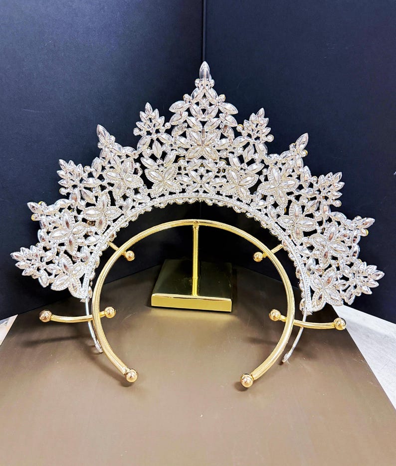 May include: A silver-toned crown with a floral design, adorned with clear crystals. The crown features a gold-colored base with a curved design and small, round accents. The piece is designed for special occasions.
