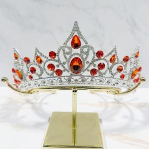 May include: A silver-toned metal tiara featuring a symmetrical, ornate scrollwork design encrusted with numerous small clear rhinestones and accented with multiple vibrant orange, teardrop-shaped and round faceted crystals throughout the piece.