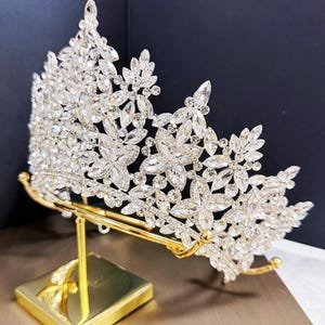 May include: A sparkling silver-toned crown with clear crystal accents, displayed on a gold-colored stand. The crown features floral and leaf designs, perfect for weddings or special events.