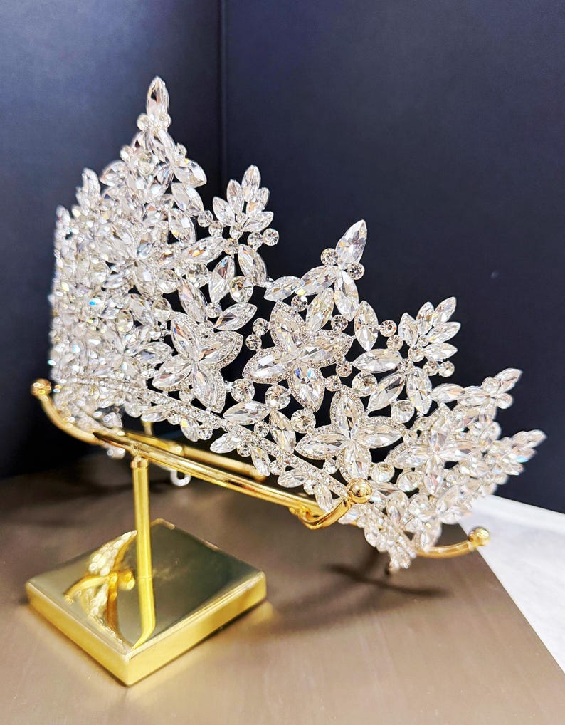 May include: A sparkling silver-toned crown with clear crystal accents, displayed on a gold-colored stand. The crown features floral and leaf designs, perfect for weddings or special events.